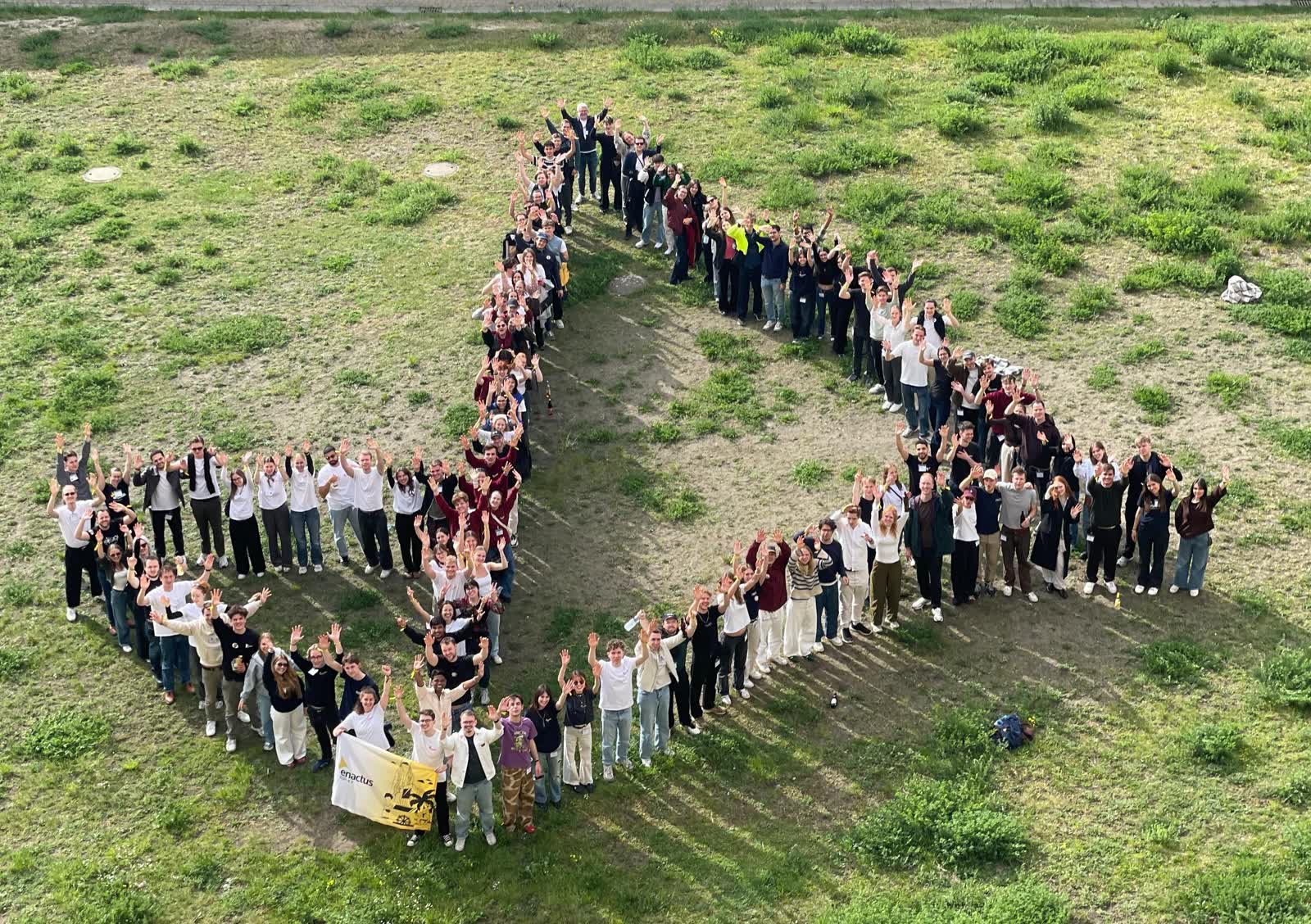 Luftaufnahme vieler Teilnehmender, die sich auf einer Wiese zu einer Formation aufgestellt haben