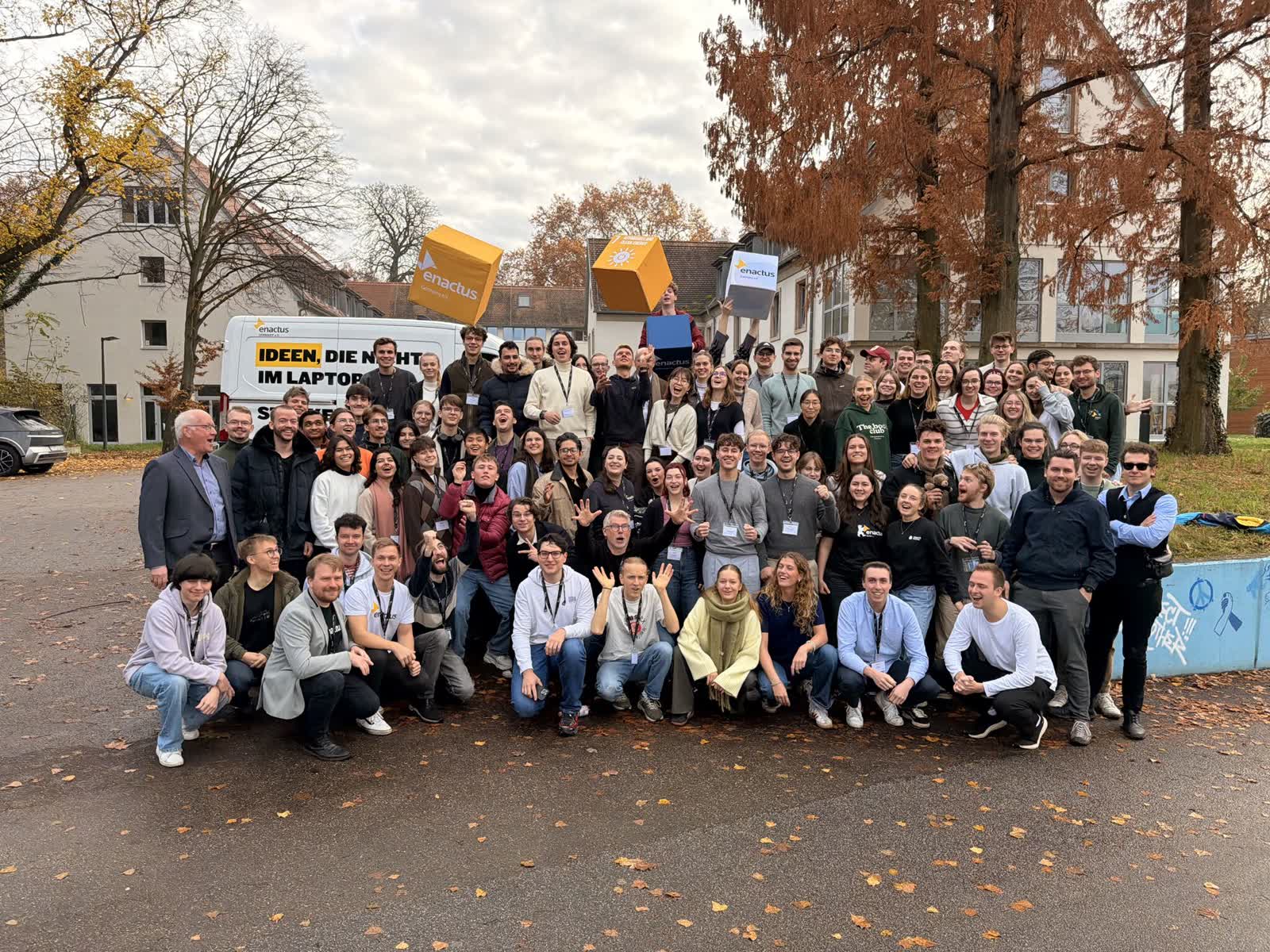 Gruppenfoto vor einem Enactus-Germany-Bus, mit hochgeworfenen bunten SDG-Boxen