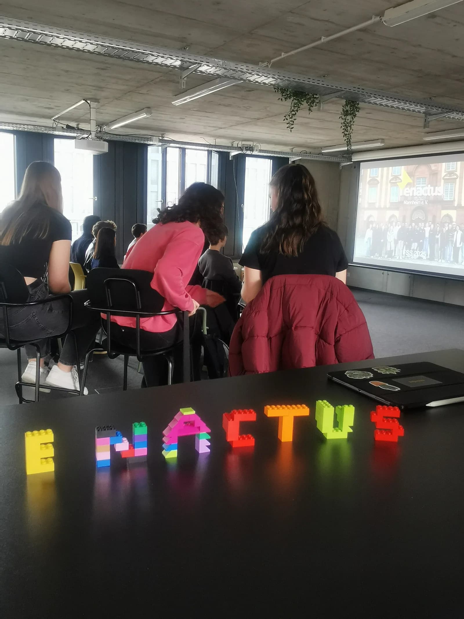 Mitglieder von hinten fotografiert, den Blick auf eine Projektionsleinwand mit dem Enactus-Logo gerichtet. Im Vordergrund liegt auf einem Tisch der Schriftzug „ENACTUS“, aus bunten Lego-Duplo-Steinen gelegt.